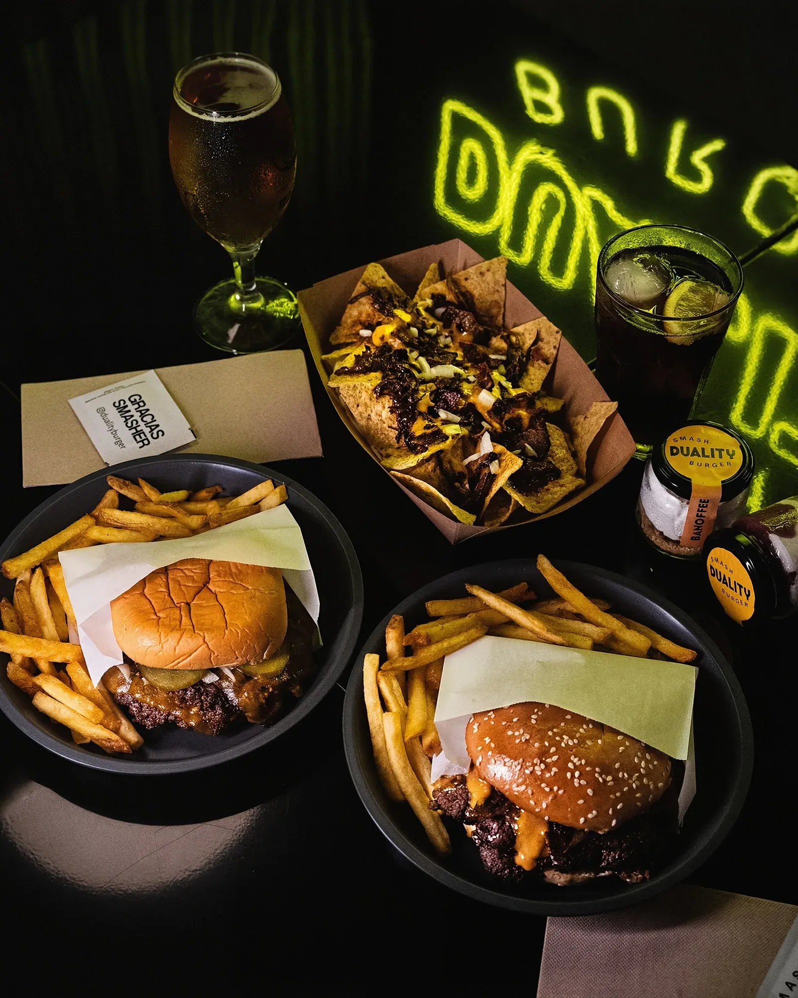Mesa vista desde arriba con dos hamburguesas y patatas en bandejas de metal, nachos, una cerveza, un refresco y una botella de Duality, con el neón al fondo