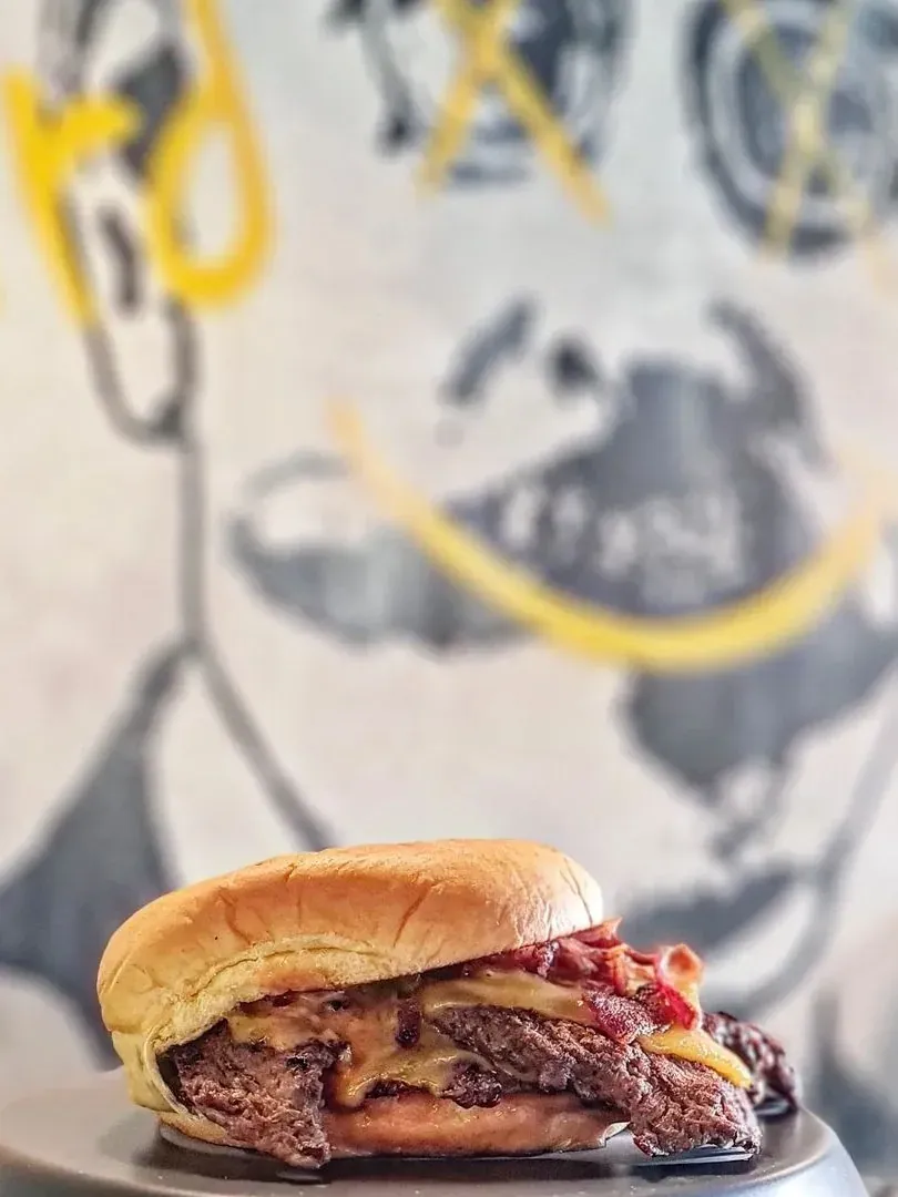 Hamburguesa con bacon y queso fundido delante del mural del local: una cara con bigote y una sonrisa pintada en amarillo
