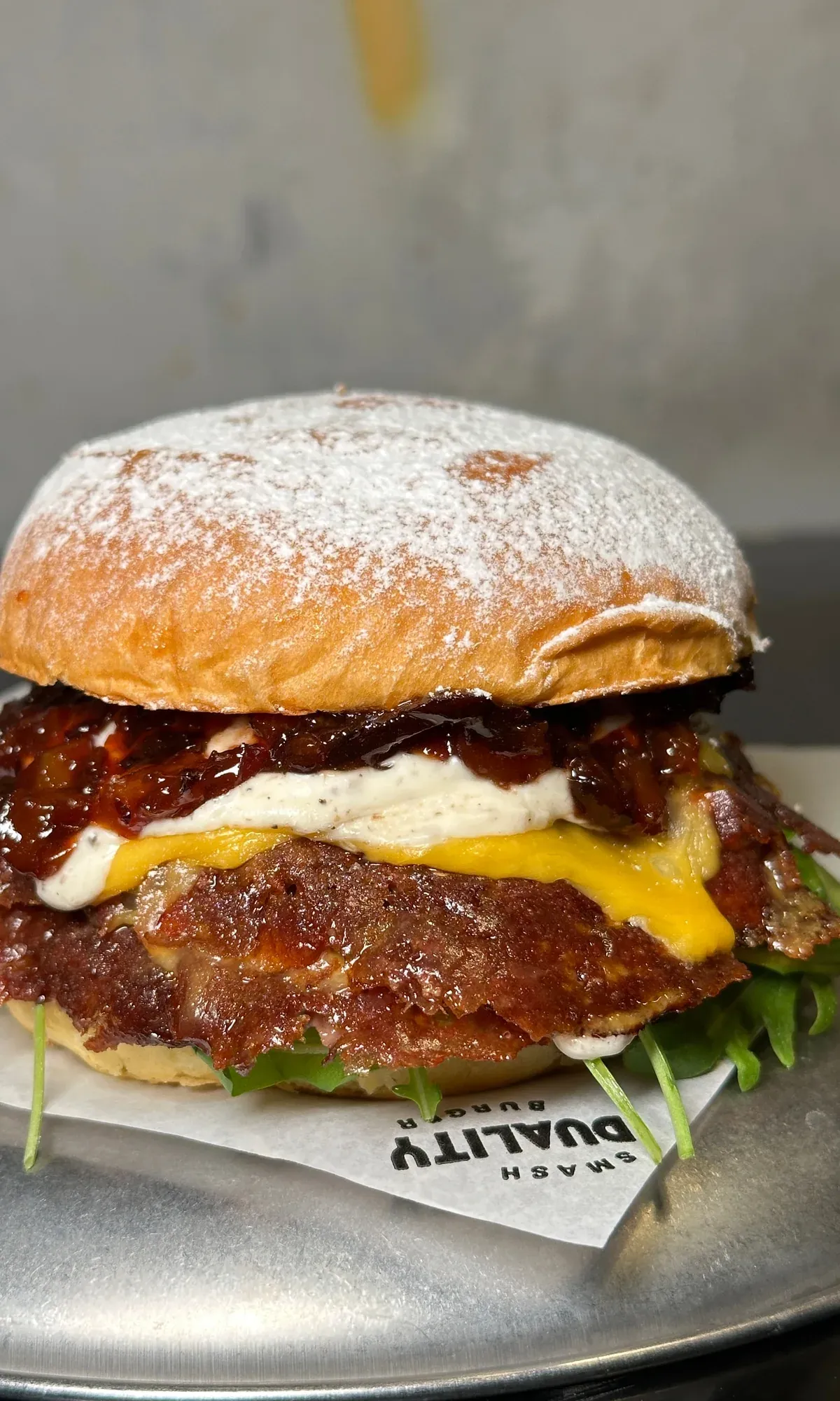 Hamburguesa de pan enharinado con cheddar, queso blanco y una capa oscura y brillante, sobre bandeja de metal