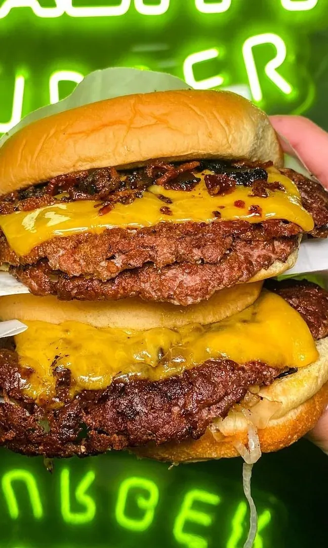 Dos manos sujetan una hamburguesa doble con cheddar fundido delante del neón verde de Duality Burger