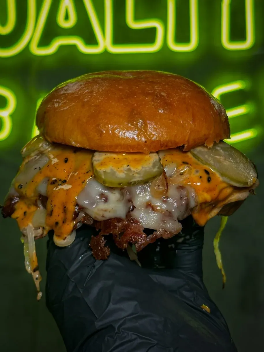 Mano con guante negro sujeta una hamburguesa con pepinillos y queso fundido ante el neón verde de Duality