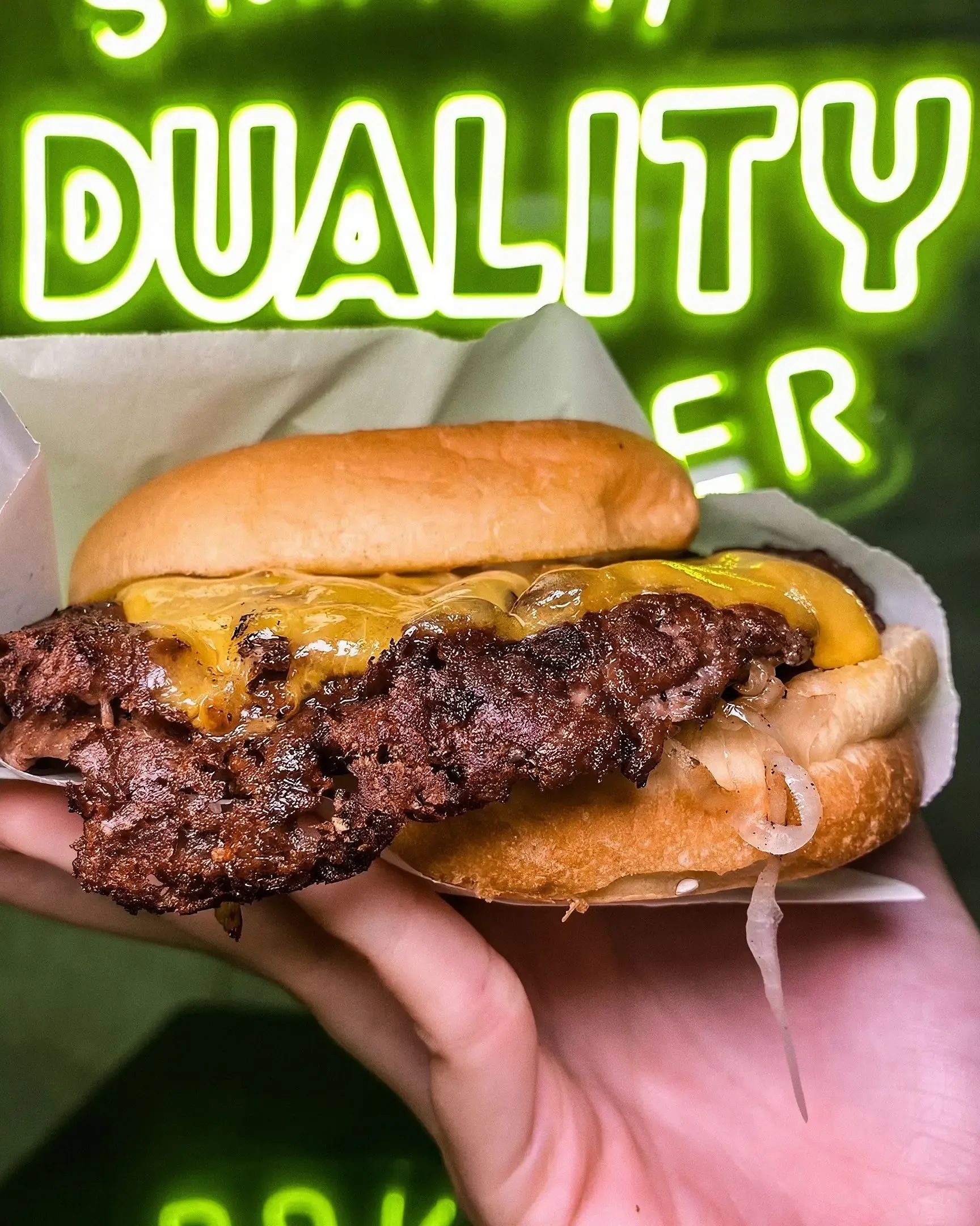Mano que sostiene una hamburguesa con cheddar en su papel, bajo el neón verde DUALITY BURGER